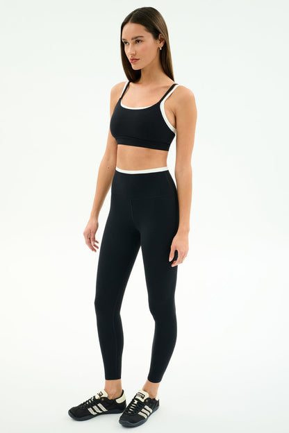 A woman stands against a white background wearing the SPLITS59 Elliah Airweight Double Layer Bra in Black/White.