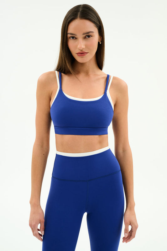 A woman poses against a plain background in the SPLITS59 Elliah Airweight Double Layer Bra in cobalt/white, styled with matching high-waisted leggings featuring white trim.