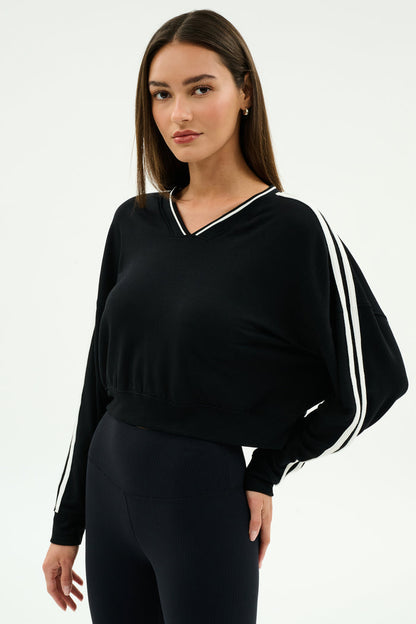 A woman stands against a plain background, wearing the SPLITS59 Ellie Fleece V Neck Sweatshirt in Black/White and black leggings designed for comfort and flexibility.