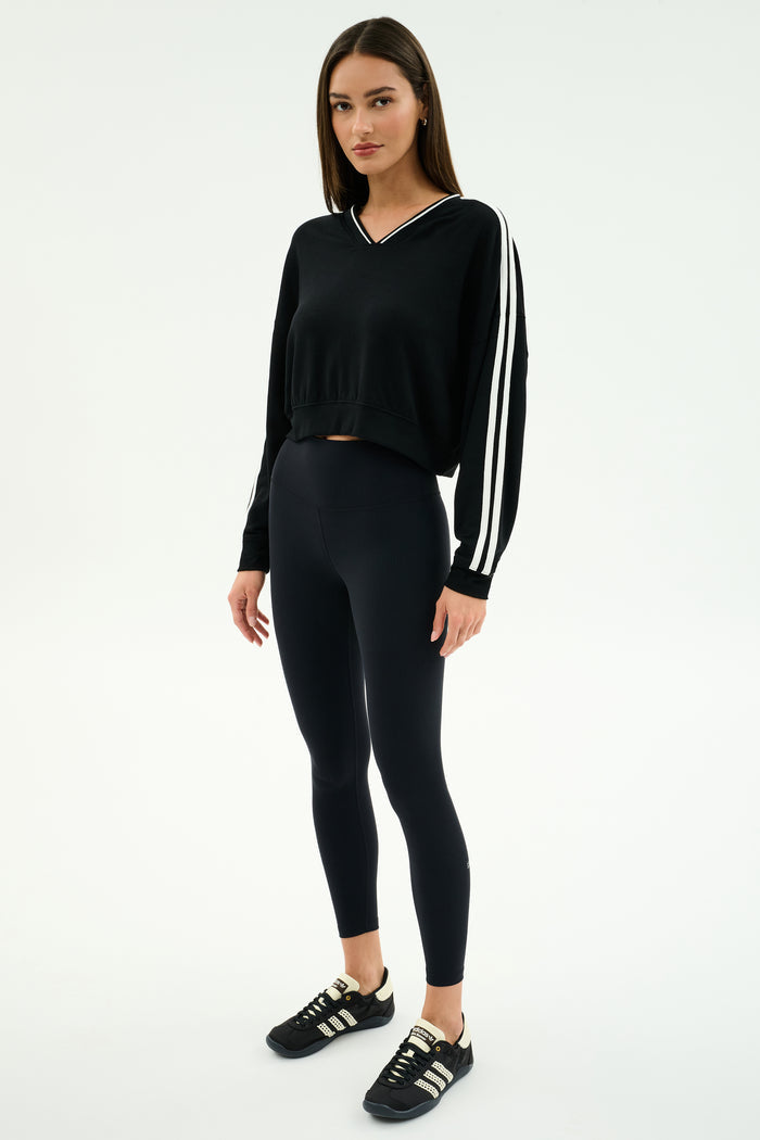 A woman stands in a studio wearing the SPLITS59 Ellie Fleece V Neck Sweatshirt in black/white, paired with black leggings and sneakers.