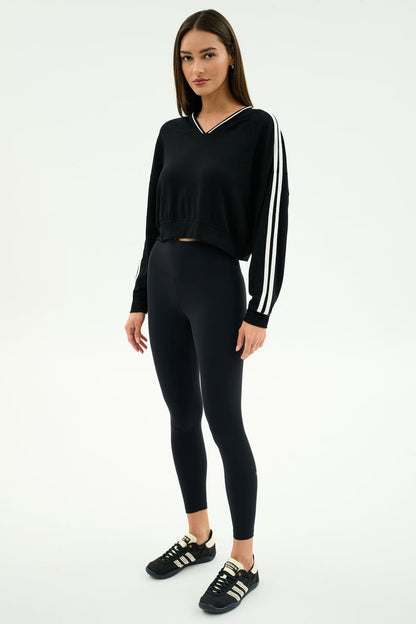 A woman stands in a studio wearing the SPLITS59 Ellie Fleece V Neck Sweatshirt in black/white, paired with black leggings and sneakers.