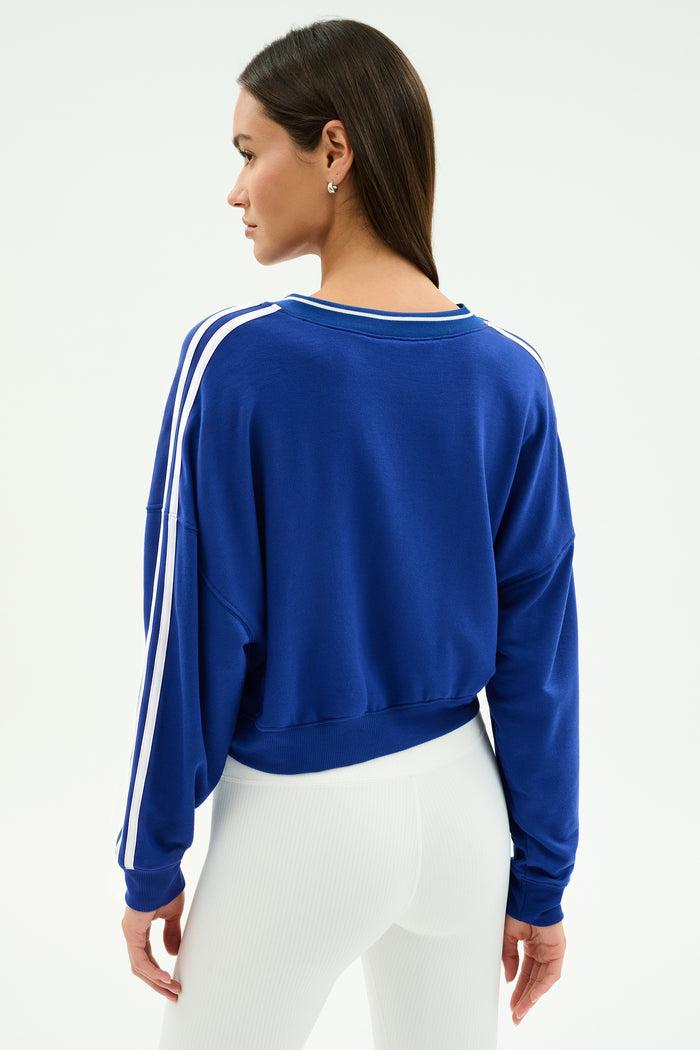 A woman stands with her back to the camera, wearing the SPLITS59 Ellie Fleece V Neck Sweatshirt in cobalt and white, paired with white high-waisted pants for an athletic look.