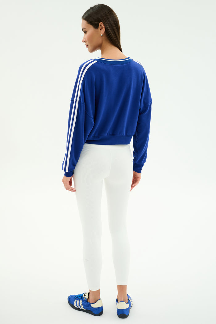 A woman faces away, wearing the SPLITS59 Ellie Fleece V Neck Sweatshirt in Cobalt/White with white leggings and blue athletic shoes, styled against a plain white background for a sporty look.