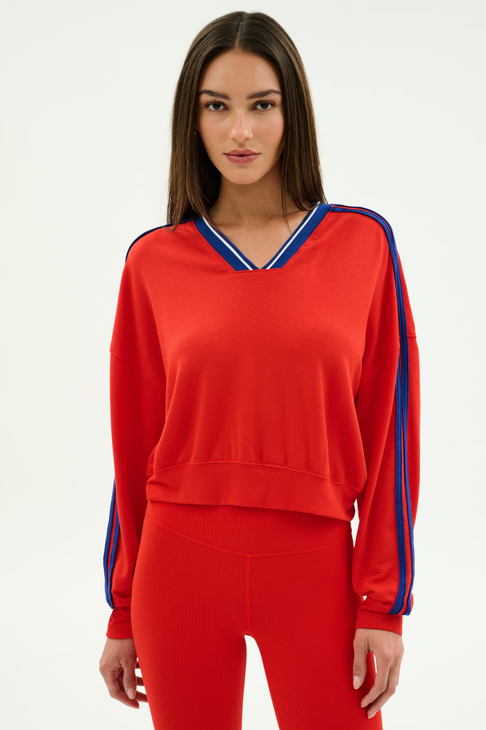 A woman stands against a plain background wearing the SPLITS59 Ellie Fleece V Neck Sweatshirt in Lotus Red/Cobalt, featuring blue sleeve stripes and crafted from a comfortable modal-spandex blend.