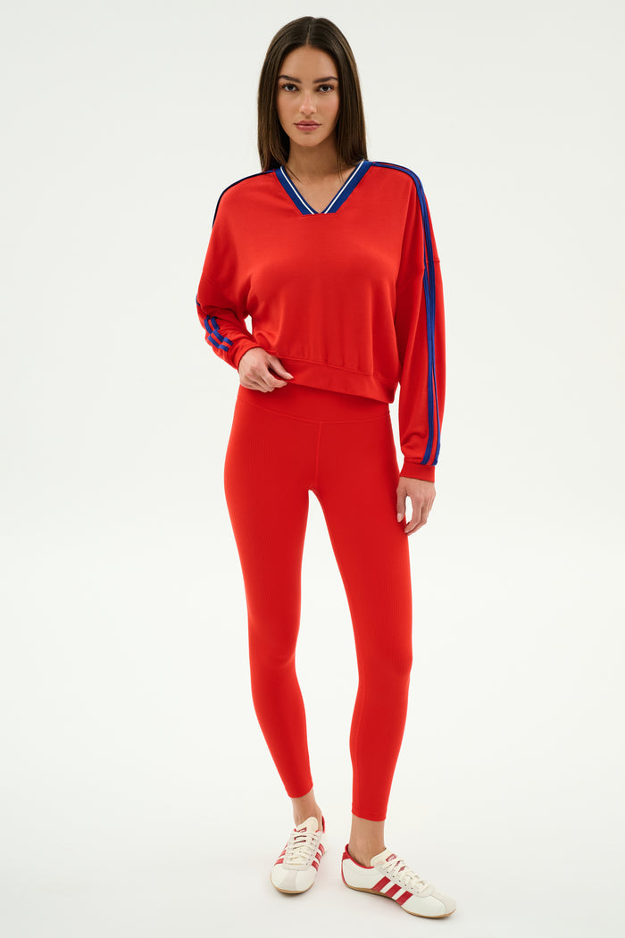 A woman wears the SPLITS59 Ellie Fleece V Neck Sweatshirt in Lotus Red/Cobalt, paired with red leggings and white sneakers with red stripes, standing against a plain white background.