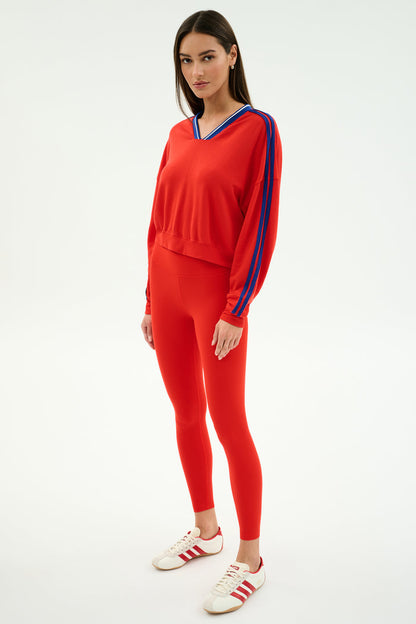 A woman wears the SPLITS59 Ellie Fleece V Neck Sweatshirt in Lotus Red/Cobalt, featuring blue sleeve stripes, paired with matching red leggings and white sneakers with red stripes.