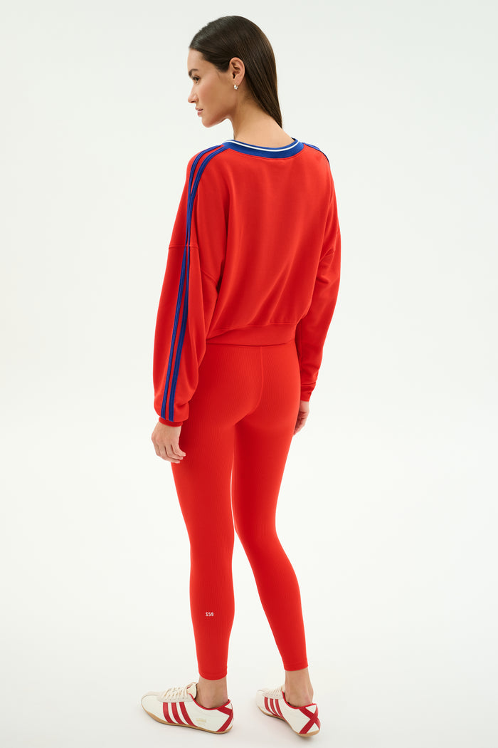A woman stands sideways against a white background, wearing the SPLITS59 Ellie Fleece V Neck Sweatshirt in Lotus Red/Cobalt with matching red leggings and white Adidas sneakers featuring red stripes.