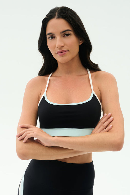 A woman with long dark hair wears the SPLITS59 Emerson Airweight Bra in Black/Ice, standing arms crossed against a plain white background—ideal for hot yoga or Pilates.