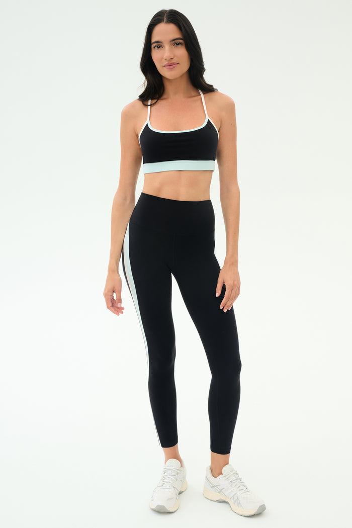A woman stands against a plain background wearing the SPLITS59 Emerson Airweight Bra in Black/Ice, paired with matching black leggings featuring a white stripe and white athletic shoes—ideal for Pilates or hot yoga.