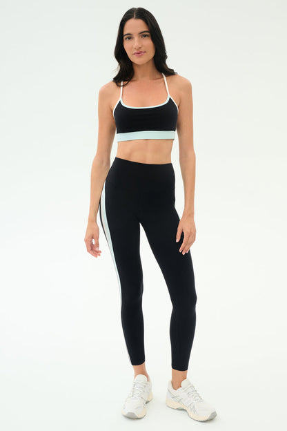 A woman stands against a plain background wearing the SPLITS59 Emerson Airweight Bra in Black/Ice, paired with matching black leggings featuring a white stripe and white athletic shoes—ideal for Pilates or hot yoga.