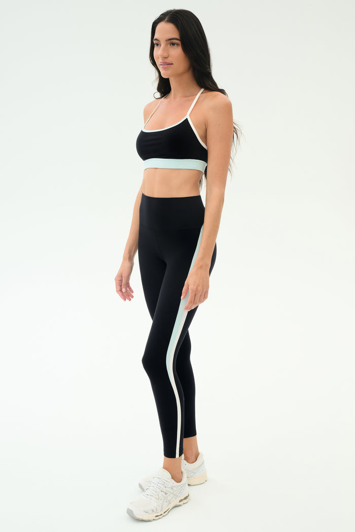 Woman in black SPLITS59 Emerson Airweight Bra - Black/Ice with white and light blue trim, standing slightly to the side against a plain white background—ideal for hot yoga or Pilates.
