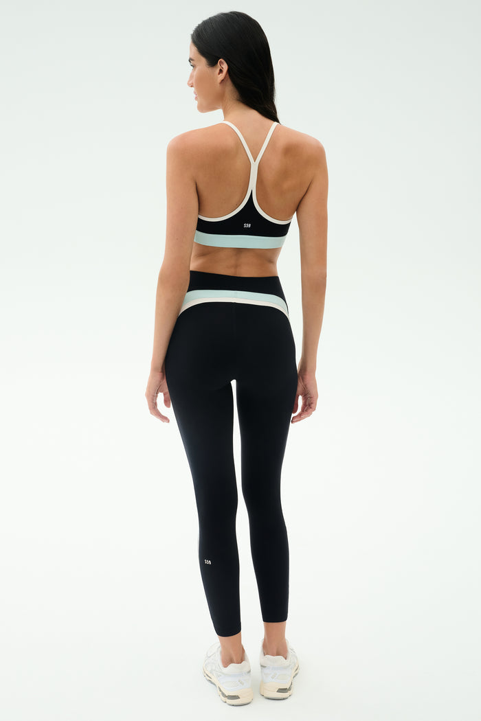 A woman faces away, wearing the SPLITS59 Emerson Airweight Bra in Black/Ice and high-waisted leggings with white accents, plus white sneakers—perfect for Pilates or hot yoga—set against a plain light background.