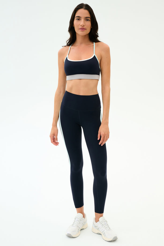 A woman stands against a plain background wearing the SPLITS59 Emerson Airweight Bra in Indigo/Dove Grey, paired with matching high-waisted leggings and white sneakers—perfect for hot yoga sessions.