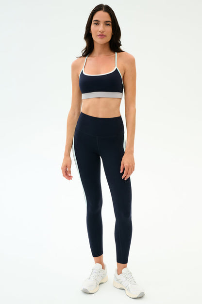 A woman stands against a plain background wearing the SPLITS59 Emerson Airweight Bra in Indigo/Dove Grey, paired with matching high-waisted leggings and white sneakers—perfect for hot yoga sessions.
