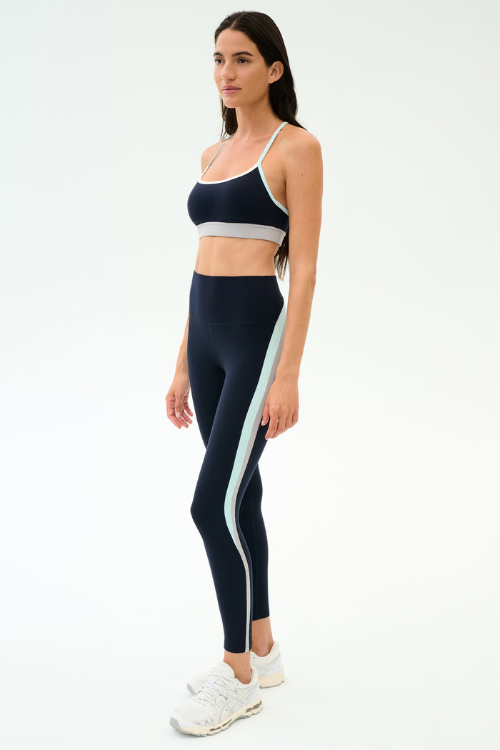 A woman wears the SPLITS59 Emerson Airweight Bra in Indigo/Dove Grey with matching high-waisted leggings, featuring a light blue stripe, styled with white sneakers against a plain light background—ideal for hot yoga.