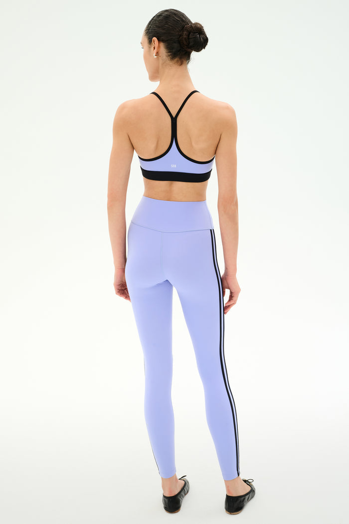 A woman faces away, wearing the SPLITS59 Emerson Airweight Bra in Purple Haze/Black and high-waisted lavender leggings with black side stripes, made from stretch fabric, set against a plain white background.
