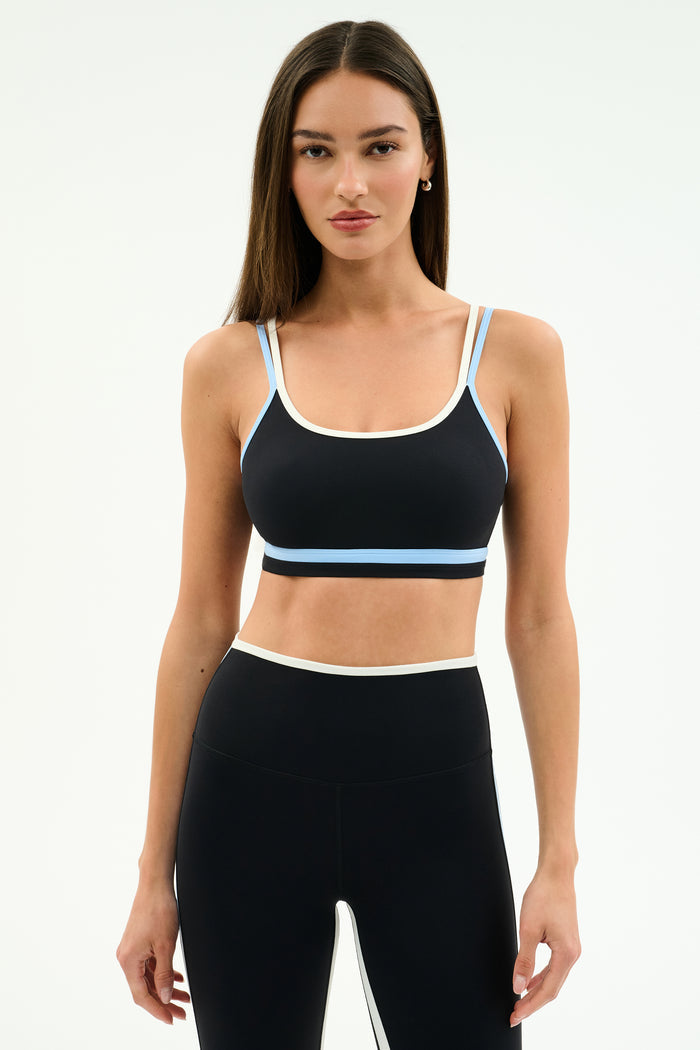 A woman stands against a plain background wearing the SPLITS59 Eva Rigor Double Strap Bra in Black/Skylight with matching high-waisted leggings.