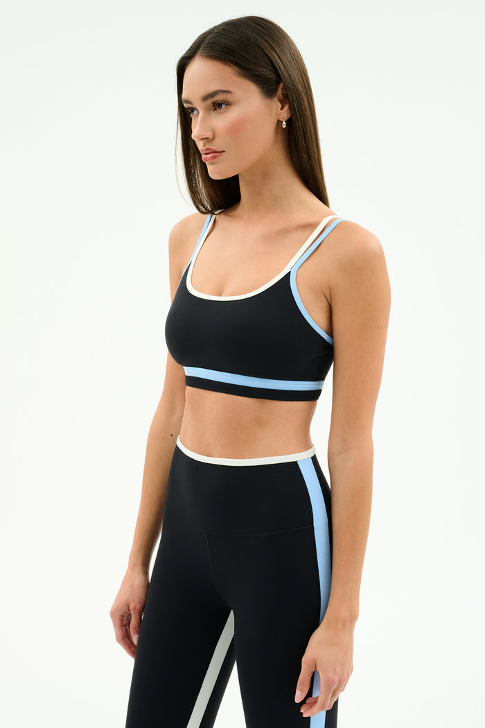 A woman wears the SPLITS59 Eva Rigor Double Strap Bra in Black/Skylight, featuring white and light blue trim, paired with high-waisted leggings, combining style, comfort, and support for any workout against a plain white background.