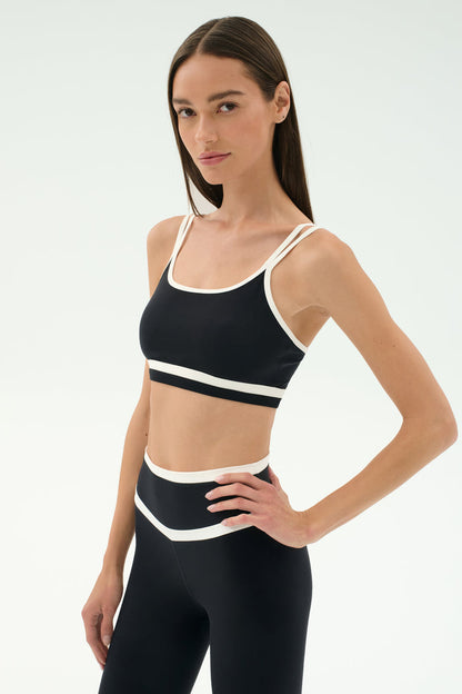 A woman wears the SPLITS59 Eva Rigor Double Strap Bra in Black/White, paired with matching high-waisted leggings, standing with one hand on her hip against a plain white background.