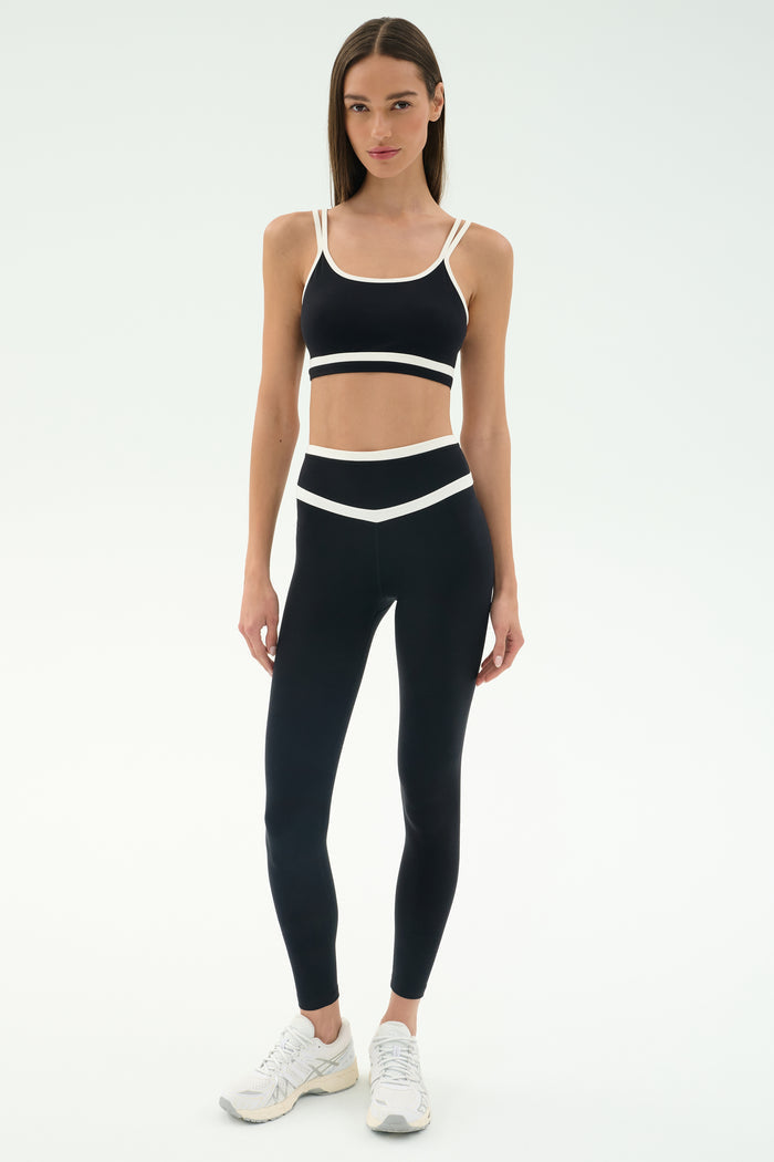 A woman faces forward in the SPLITS59 Eva Rigor Double Strap Bra - Black/White with matching high-waisted leggings and white athletic shoes, ready for gym workouts against a plain light background.