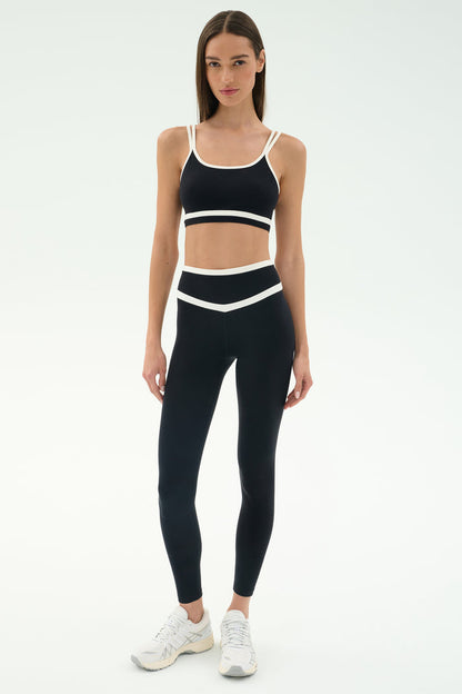 A woman faces forward in the SPLITS59 Eva Rigor Double Strap Bra - Black/White with matching high-waisted leggings and white athletic shoes, ready for gym workouts against a plain light background.