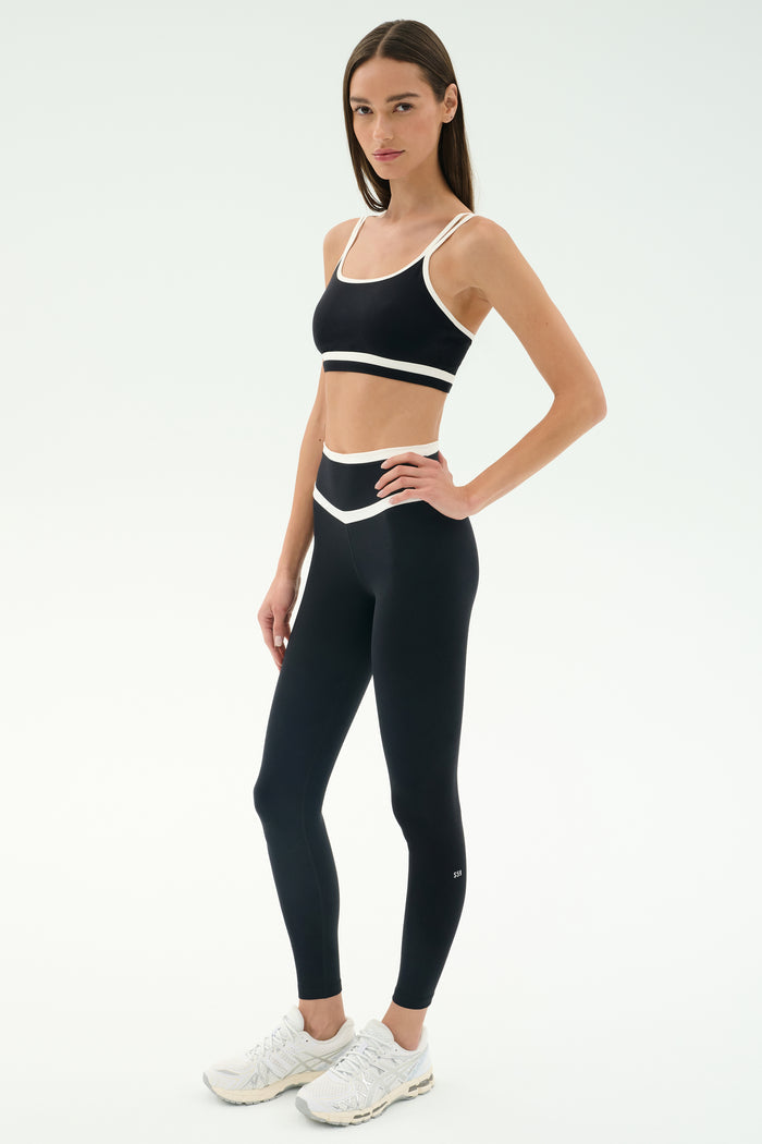 A woman stands in a studio wearing the SPLITS59 Eva Rigor Double Strap Bra in Black/White and matching leggings, paired with white sneakers—an ideal look for gym workouts.
