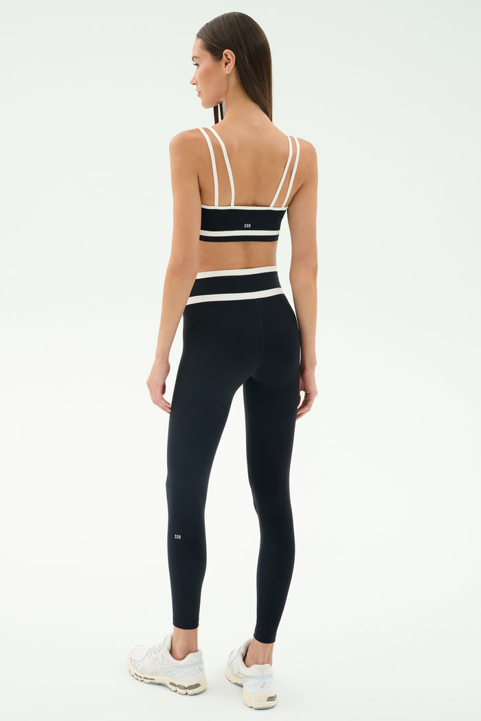 A woman faces away against a plain light background, wearing the SPLITS59 Eva Rigor Double Strap Bra in black/white, paired with matching high-waisted leggings and white sneakers, ready for a gym workout.