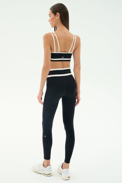 A woman faces away against a plain light background, wearing the SPLITS59 Eva Rigor Double Strap Bra in black/white, paired with matching high-waisted leggings and white sneakers, ready for a gym workout.