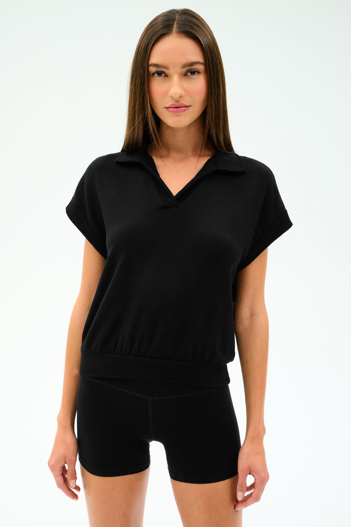 A woman wearing the SPLITS59 Evie Fleece Sleeveless Polo in black, paired with black shorts, stands against a plain light background. The look features sporty style and comfort.