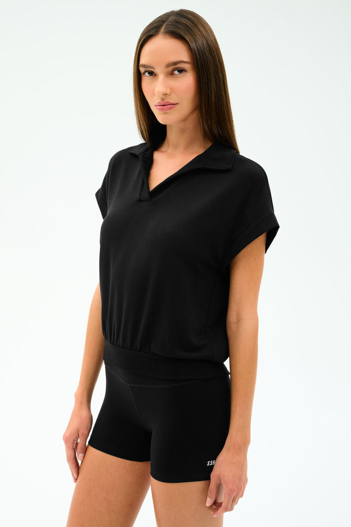 A woman with straight brown hair wears the SPLITS59 Evie Fleece Sleeveless Polo in black, paired with black fitted shorts, standing against a plain light background.
