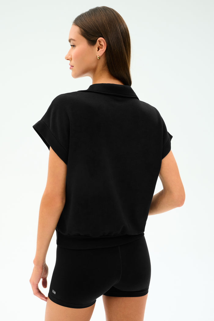 A woman faces away, wearing the SPLITS59 Evie Fleece Sleeveless Polo in black, paired with matching shorts, set against a plain light background.