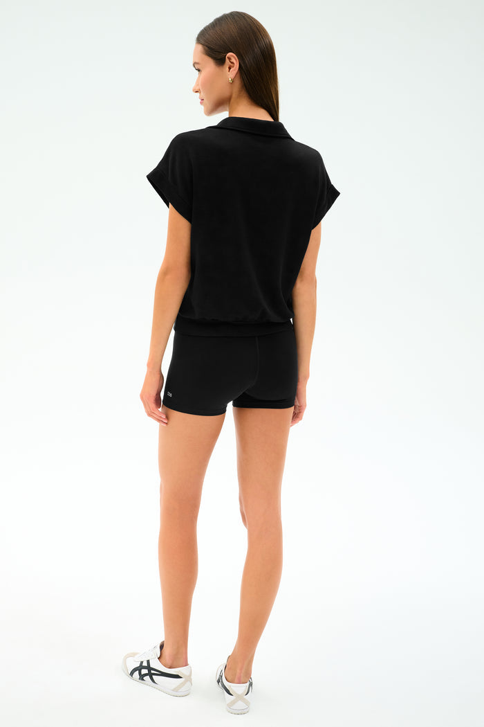 A woman faces away from the camera against a plain background, wearing the SPLITS59 Evie Fleece Sleeveless Polo in black paired with black shorts and white sneakers.