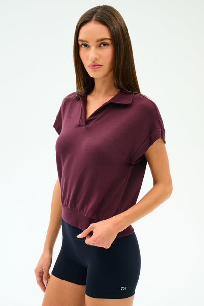 A woman with straight brown hair wears the SPLITS59 Evie Fleece Sleeveless Polo in Dark Cherry and black fitted spandex shorts against a plain light background.