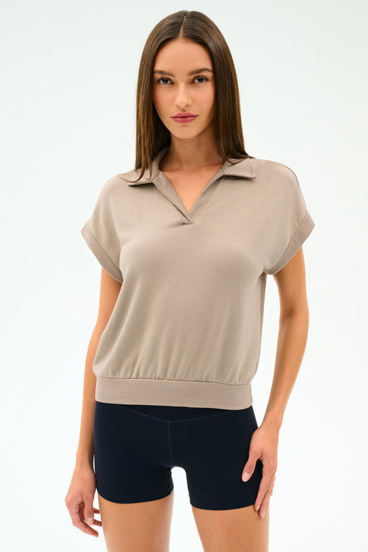 A woman poses against a plain background wearing the SPLITS59 Evie Fleece Sleeveless Polo in Latte and black fitted stretch shorts.