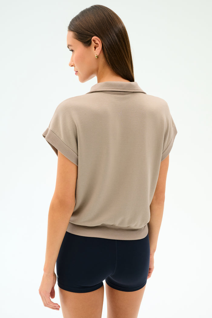 A woman stands with her back turned, wearing the SPLITS59 Evie Fleece Sleeveless Polo in Latte against a plain light background.