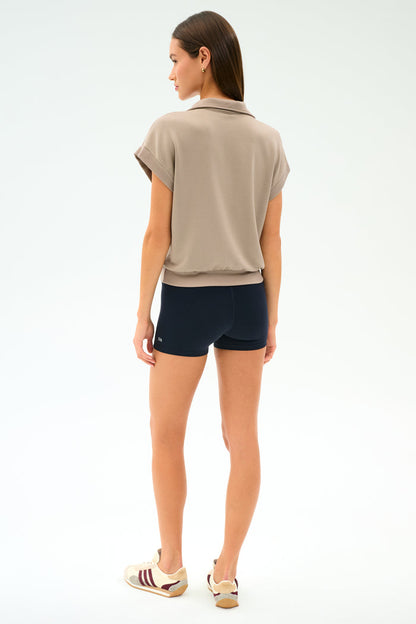 A woman stands facing away, wearing the SPLITS59 Evie Fleece Sleeveless Polo in Latte, black shorts, and beige sneakers with burgundy stripes against a plain white background.
