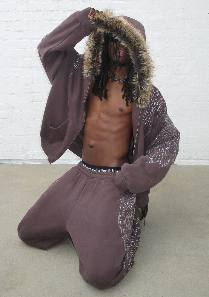 Model kneeling in a brown hoodie with detachable faux fur hood, showcasing rhinestone wing design on the sleeve.
