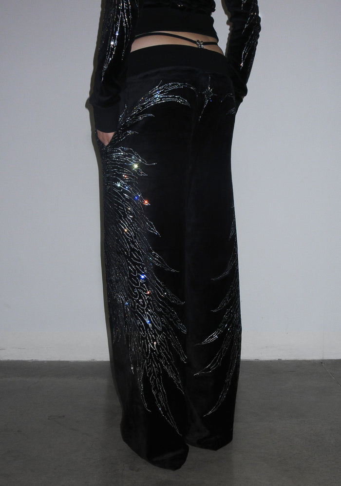Model wearing black fallen rhinestone velour sweatpants with large rhinestone wing design at the back.