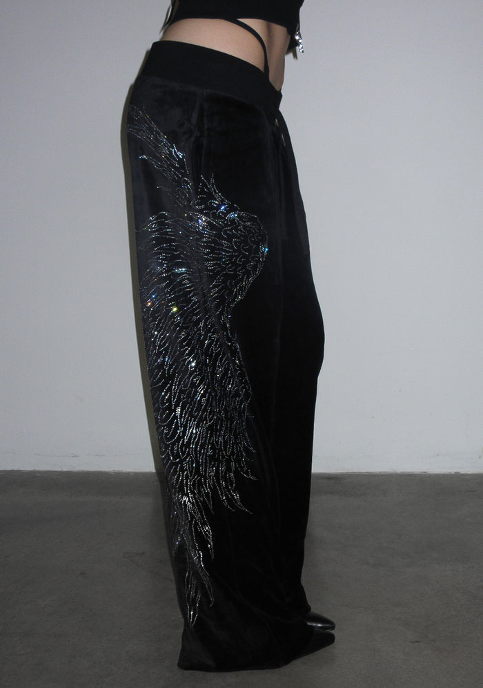 Model wearing black fallen rhinestone velour sweatpants featuring a large rhinestone wing design, side view.