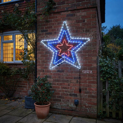 A large five-pointed star-shaped LED light decoration mounted on a red brick wall at dusk, with white outer rings, a blue middle ring, and a red center ring. A lit window, potted plants, and garden foliage are visible around it.