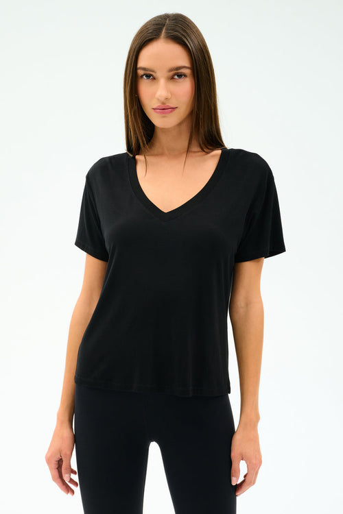 A woman faces forward, wearing the SPLITS59 Georgia Jersey V-Neck in black—crafted from a soft viscose-spandex blend—paired with black pants, set against a white background.