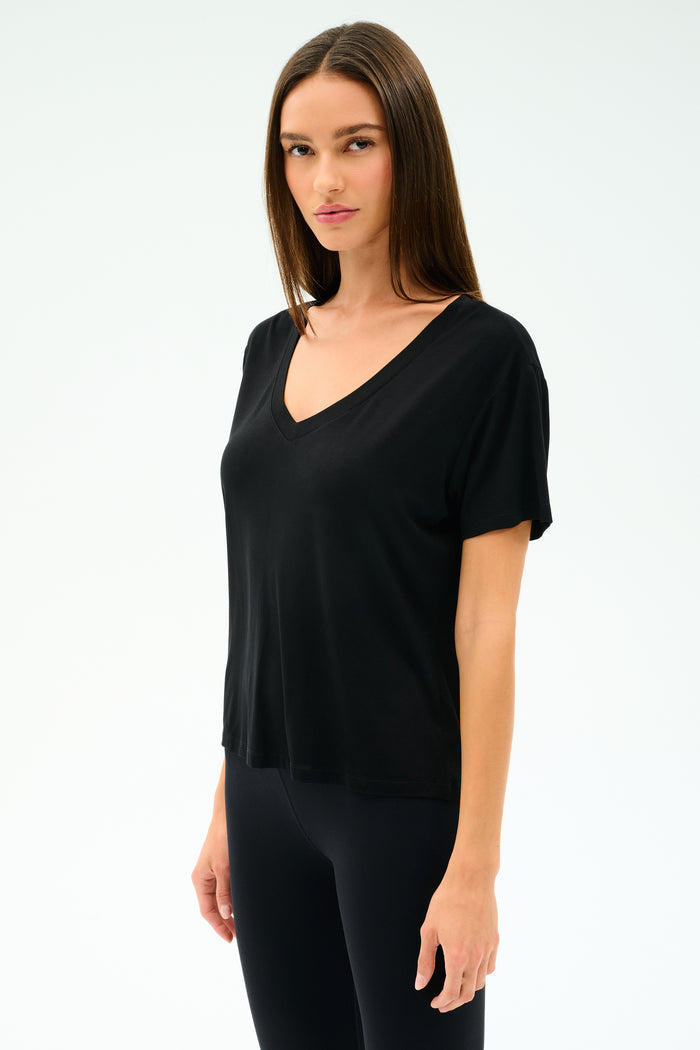 A woman with long straight hair wears the SPLITS59 Georgia Jersey V-Neck in black, styled with black leggings, standing against a plain light background.