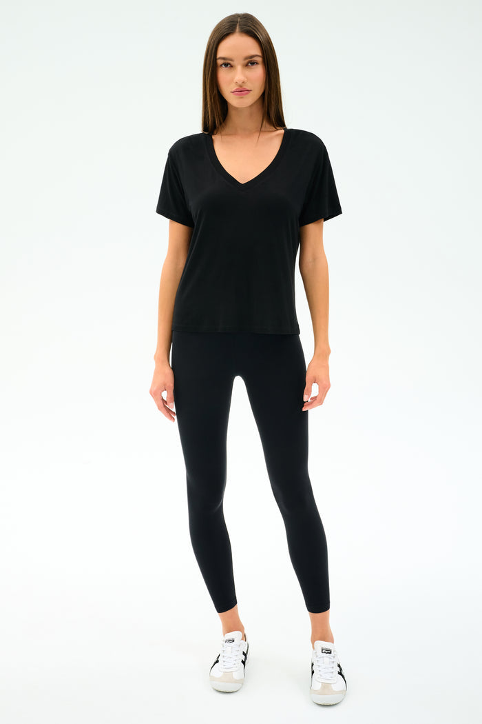 A woman faces forward, wearing the SPLITS59 Georgia Jersey V-Neck in black—crafted from soft viscose and spandex—with black leggings and white sneakers, set against a plain white background.