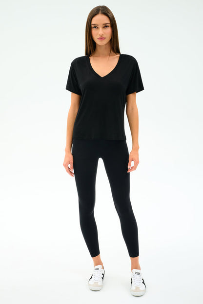 A woman faces forward, wearing the SPLITS59 Georgia Jersey V-Neck in black—crafted from soft viscose and spandex—with black leggings and white sneakers, set against a plain white background.
