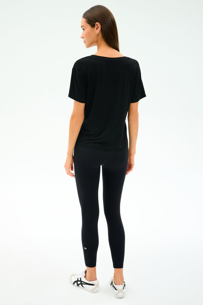 A woman faces away, wearing the SPLITS59 Georgia Jersey V-Neck in black, paired with black leggings and white sneakers against a light background.