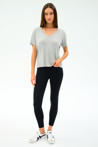 A woman faces forward in a SPLITS59 Georgia Jersey V-Neck in Heather Grey, paired with black spandex leggings and white sneakers, set against a plain white background.