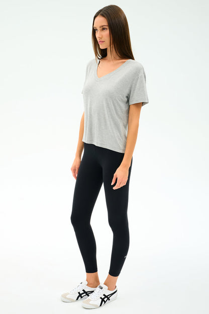 A woman stands against a plain background, wearing the SPLITS59 Georgia Jersey V-Neck in Heather Grey, paired with black leggings and white sneakers featuring black stripes.