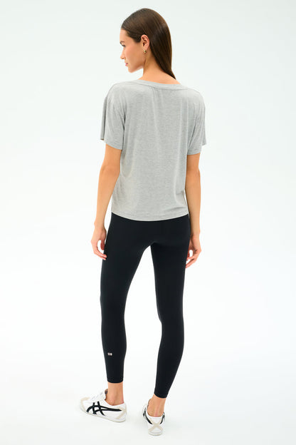 A woman stands facing away from the camera, wearing the SPLITS59 Georgia Jersey V-Neck in Heather Grey, black spandex leggings, and white sneakers against a plain white background.
