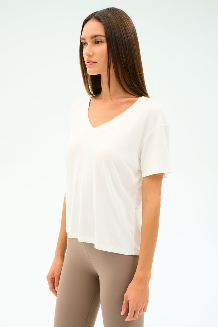 A woman stands against a plain light background wearing the SPLITS59 Georgia Jersey V-Neck in white, a loose-fitting T-shirt crafted from a soft viscose blend, paired with beige leggings.