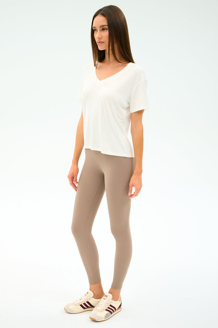 A woman stands against a plain background wearing the SPLITS59 Georgia Jersey V-Neck in white, paired with beige leggings and beige sneakers featuring brown stripes.